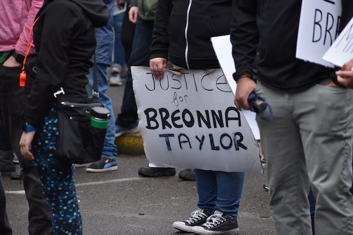 The legs and torsos of people gathered for a protest are pictured. At knee height, person holds a protest sign that says 